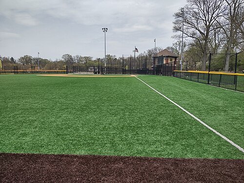 A synthetic turf ballfield in Branch Brook Park, bare trees and a fieldhouse with a cupola beyond the outfield fence.