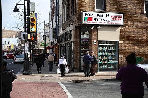A brick corner building with the Portugalia Sales import shop sign, a window display of decorative bottles, pedestrians on the sidewalk.