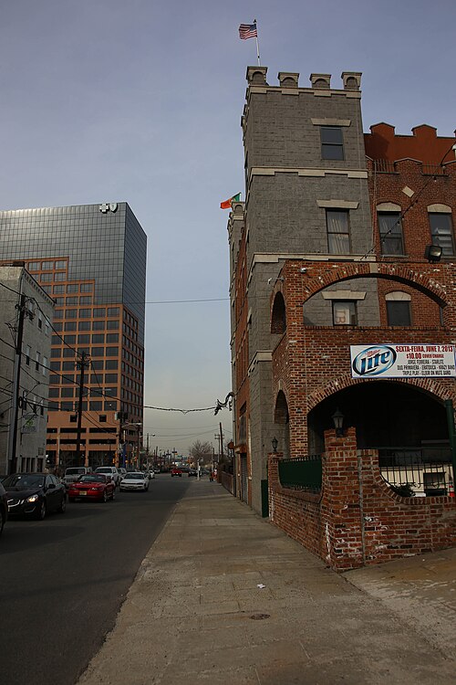 A castellated brick and stone corner building with turrets and an American flag, downtown Newark's office towers rising behind it along the street.