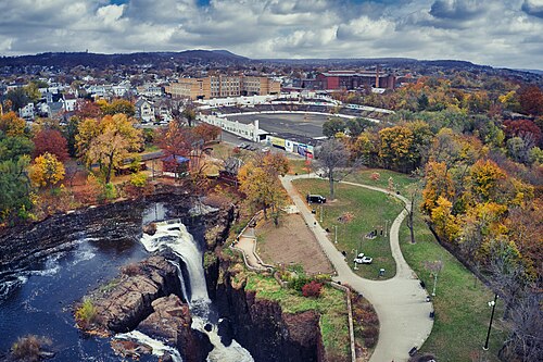 An aerial view of the falls, the paved overlook path along the gorge rim and Hinchliffe Stadium and downtown Paterson's mill blocks behind the trees.