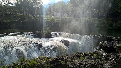 A lower, wider spillway upstream of the main falls, sunlight catching the spray and cars parked along the road behind the trees.