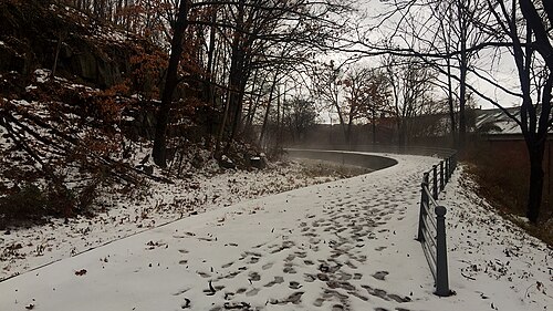 A snow covered paved path curving along the Upper Raceway, a stone retaining wall and wooded slope on one side.