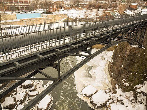A steel pipe bridge crossing the gorge in snow, a large pipeline running alongside its walkway above the frozen river.
