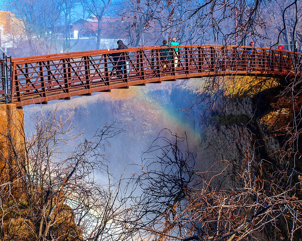 The steel footbridge over the gorge, its wood decked walkway crossing above rising mist and a rainbow, visitors leaning on the rail.