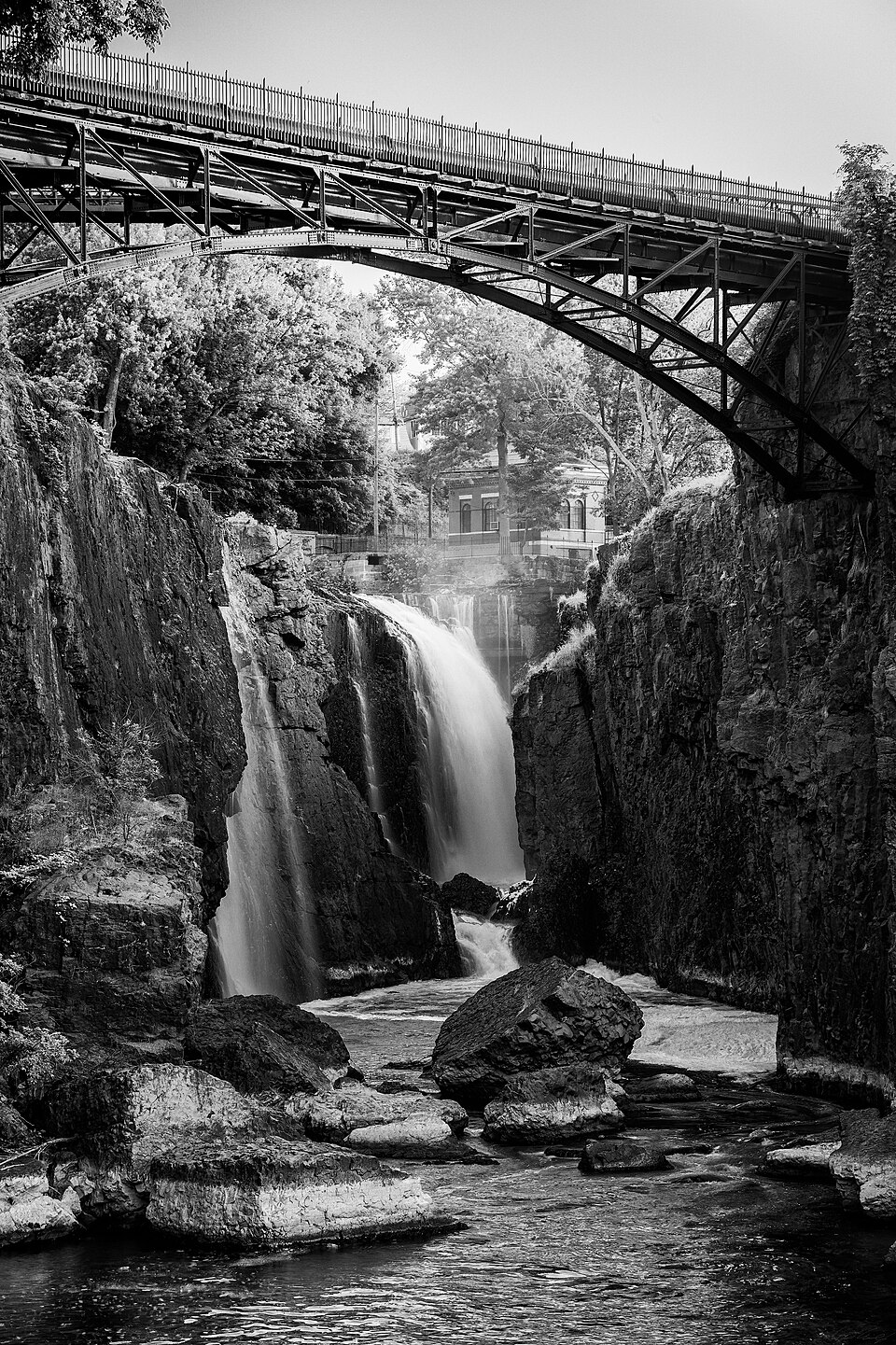 A monochrome view from the gorge rim near the falls, the arch bridge crossing overhead and a building glimpsed through the trees beyond.