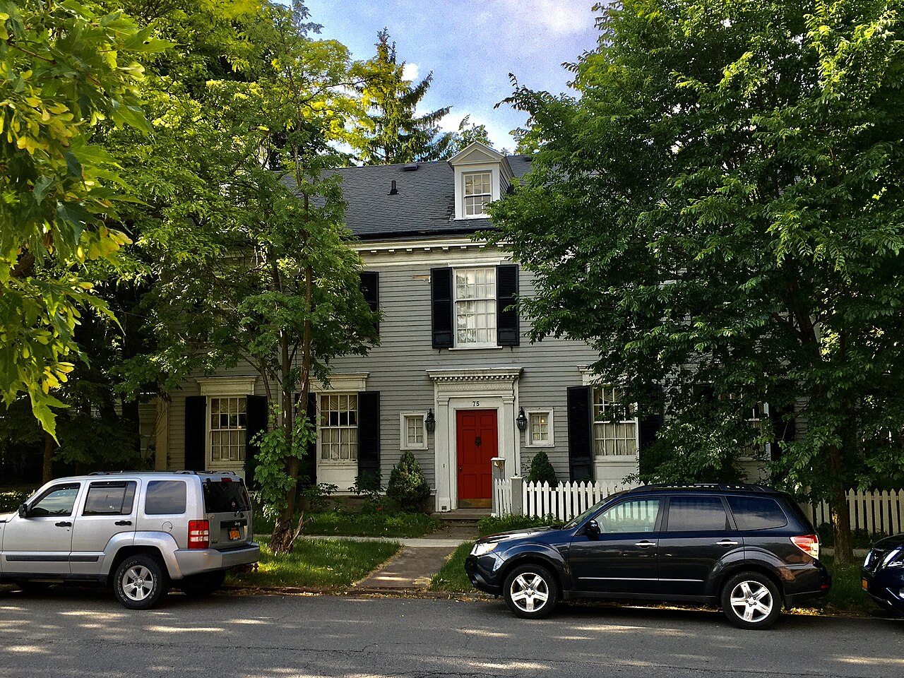 A two storey grey clapboard house with black shutters, a red front door and a white picket fence.