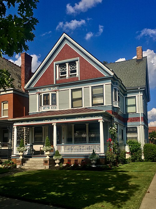 A two and a half storey Queen Anne house with grey clapboard siding, a cross gable and a full width porch.