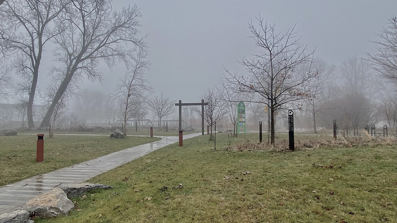 A paved riverside trail with bare trees and wooden posts, fog settling over the ward beside Ohio Street.