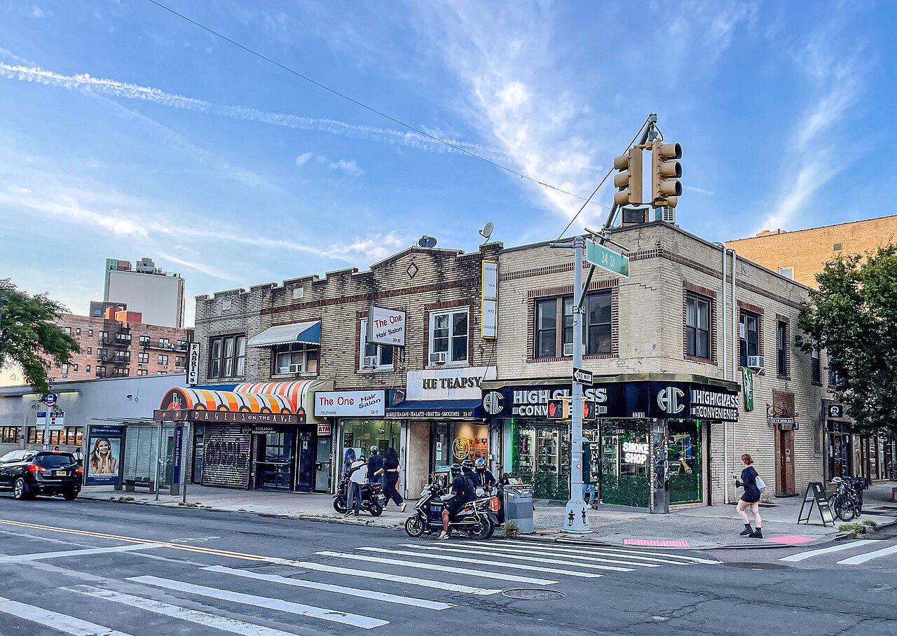 A lively 30th Avenue corner, bagel shop awning and storefronts with pedestrians.
