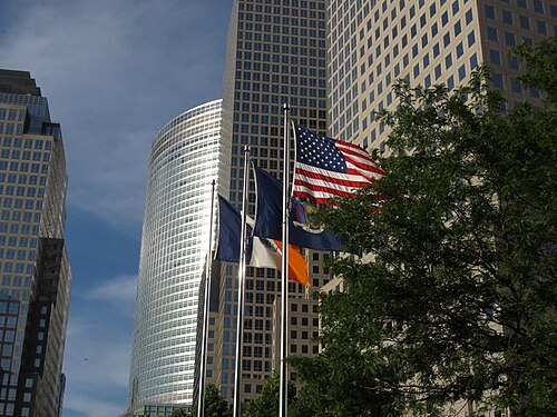 American, New York State and New York City flags flying in front of Brookfield Place's granite and curved glass office towers, framed by tree branches.