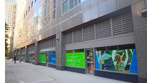Ground floor shopfronts under a grey granite office tower along South End Avenue, storefront glass reflecting the block across the street.