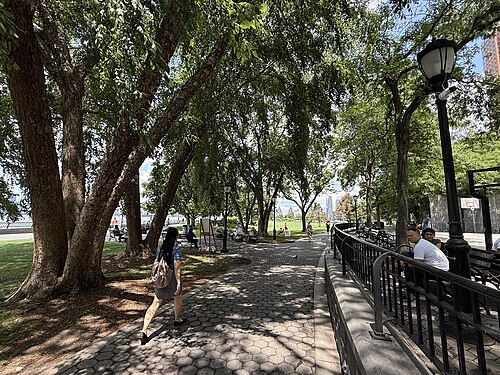 A brick paved path under a row of trees in Rockefeller Park, benches and an iron lamppost along the rail and the Jersey City skyline visible through the leaves.