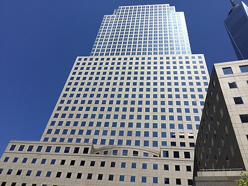 A glass and pale stone office tower rising past a darker granite neighbor, with One World Trade Center's spire visible behind it.