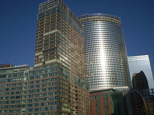 An orange brick apartment tower with glass balconies at the neighborhood's north end, a curved glass office tower rising just outside the neighborhood across West Street.