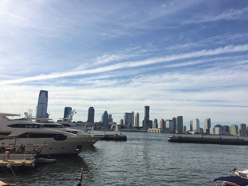 Motor yachts moored along North Cove Marina's dock, masts and flags flying, with the Jersey City skyline across calm harbor water.