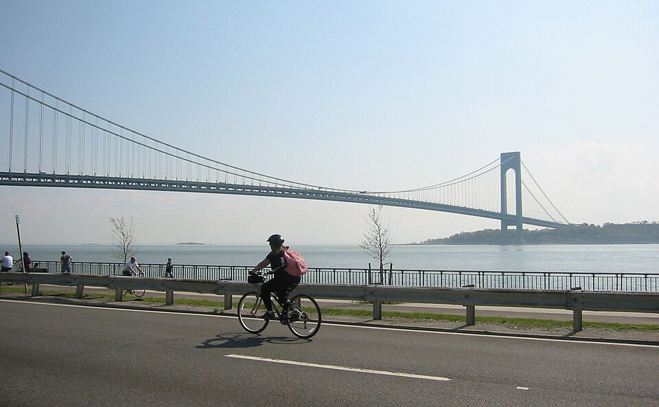 The Verrazzano-Narrows Bridge from the paved path along the water in Bay Ridge, a cyclist going past on the roadway and Staten Island on the far side of the Narrows.