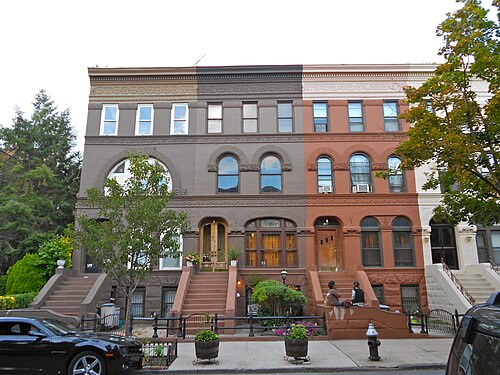 Three brownstones on the 200 block of Decatur Street, each stoop painted a different colour, two people sitting on the steps of the one on the right.