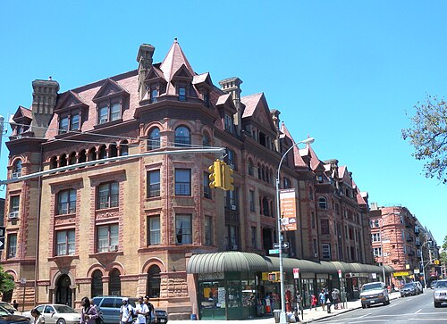 The Alhambra Apartments looking north along Nostrand Avenue across Macon Street, red brick and brownstone with dormers and a corner turret over a run of shop awnings.