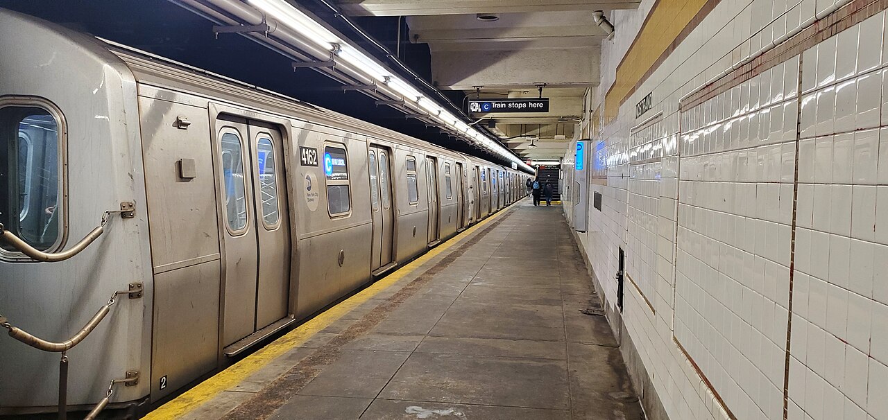 The Brooklyn bound local platform at Nostrand Avenue on the A and C, white tiled wall along the right, a train standing at the platform edge.