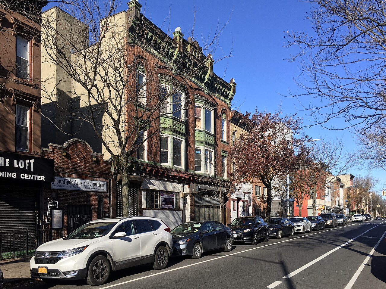 The west side of Tompkins Avenue between Madison and Monroe Streets on a clear winter afternoon, a storefront church sign on the low brick building at the left and a taller house beside it with green painted bays and a heavy cornice, cars parked the length of the block.