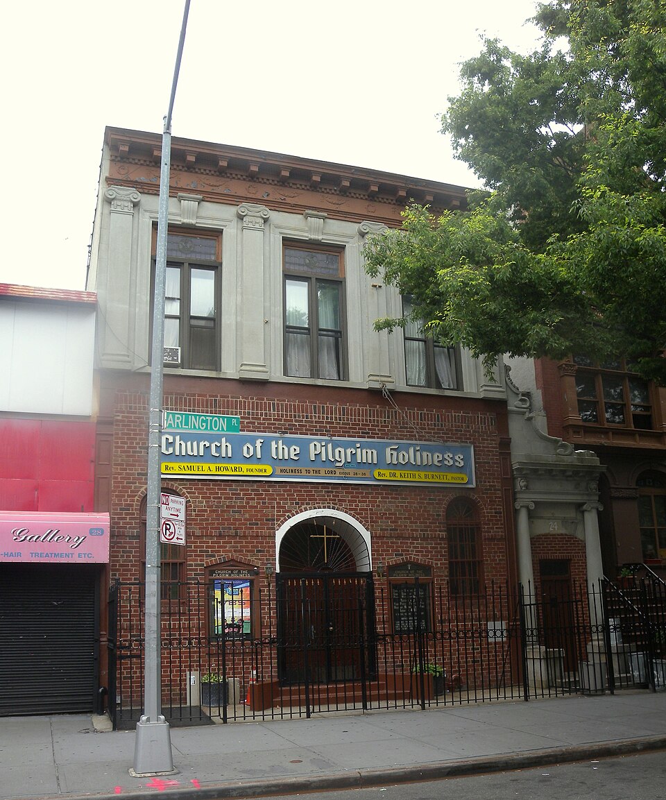 The Church of the Pilgrim Holiness at 24 Arlington Place, a two storey brick front with a painted sign across it and an iron fence along the sidewalk, the ARLINGTON PL street sign on the pole in front.