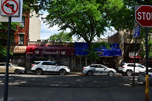 Biancardi's butcher shop and Madonia Brothers Bakery side by side at 2350 and 2348 Arthur Avenue, red and blue awnings under the street trees.