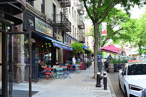 Bistro tables on the sidewalk outside Parisienne Cafe at 2339 Arthur Avenue, Enzo's next door and planters along the parking lane.