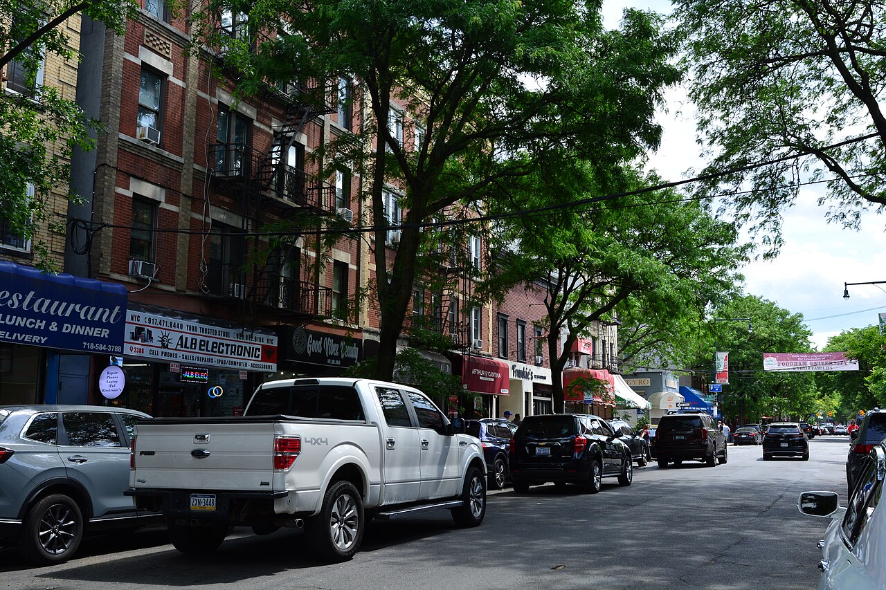 Arthur Avenue between East 186th and East 187th Streets, shopfronts and awnings under brick apartment houses with fire escapes, a banner over the roadway in the distance.