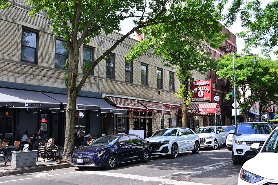 The east side of Arthur Avenue below East 186th Street, cafe tables on the sidewalk and the Pasquale's Rigoletto sign on the two storey building.