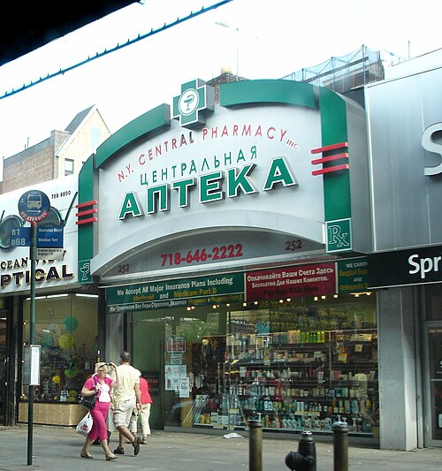 The front of the N.Y. Central Pharmacy on Brighton Beach Avenue, the Russian word for pharmacy in green letters two feet high and the English name small above it, with an optical shop next door and the window packed with stock.