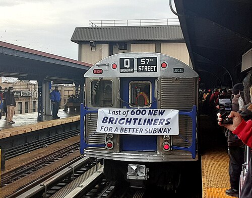 A stainless steel R32 train signed for the Q standing at the Brighton Beach elevated station, with a banner across its front and a crowd photographing it from the platform.