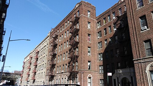 Six storey brick apartment houses on Brighton 7th Street with red fire escapes down the fronts, arched windows at street level and a corner tower at the end of the row.