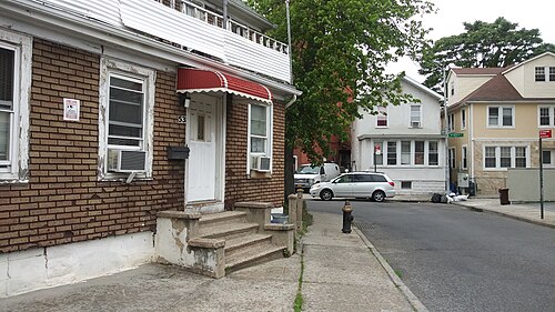 A narrow lane between Brighton 1st and Brighton 2nd Streets, small brick and wood houses with concrete stoops, window air conditioners and a red striped awning over one door.