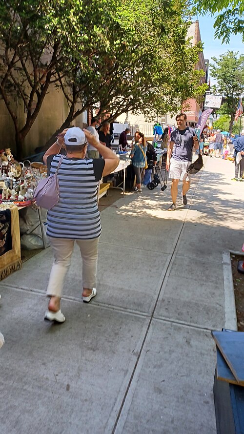 Folding tables of second hand china and jewelry set out against a blank wall at the Brighton 4th Street flea market, with racks of clothes further along and a beauty salon sign down the block.