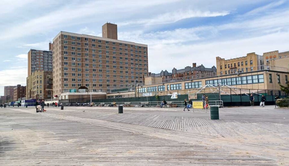 The Riegelmann Boardwalk at Brighton 6th Street, sand blown across the planks, the glassed in restaurant fronts along the right and an eleven storey apartment block behind them.
