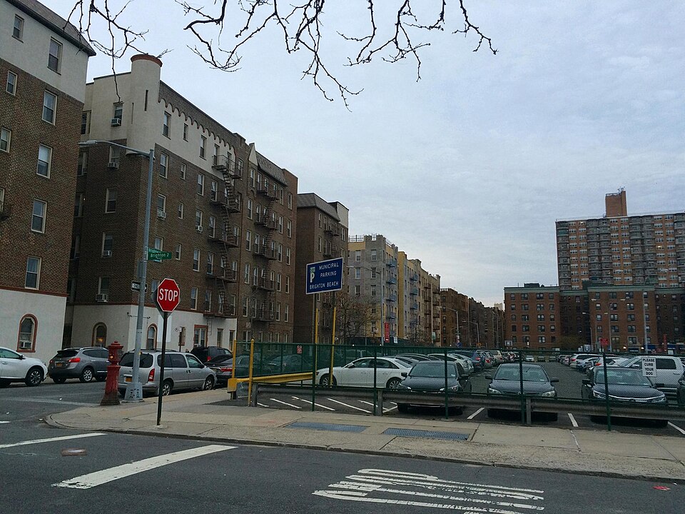 Six storey brick apartment houses along the north side of Brightwater Court, seen from the corner of Brighton 2nd Street, with a municipal parking lot behind a green fence in the middle of the block.