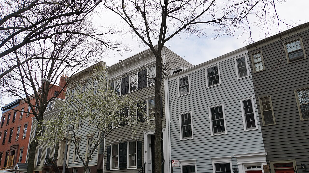 Wood-frame houses on Middagh Street in early spring, clapboard and shingle fronts painted grey and white, black shutters on one and a tree in white blossom in front of it.