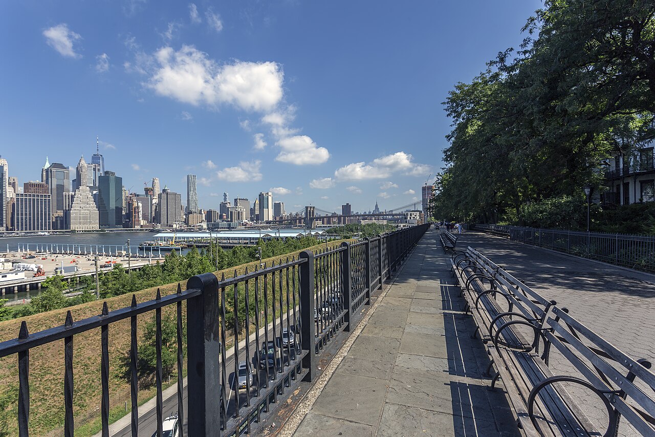 The Promenade looking north on a clear afternoon, benches along the right and an iron railing along the left, with the expressway lanes visible through it and Lower Manhattan across the water.