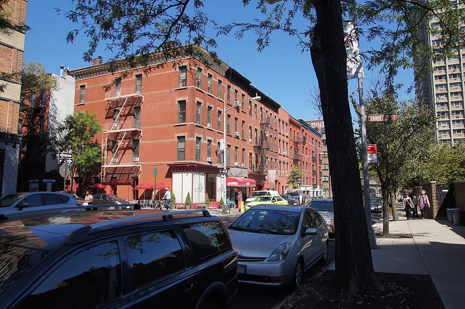 The Cranberry Street sign on a corner pole near the east end of the block, a red brick building behind it with fire escapes on the front and awnings over the shops at the sidewalk.