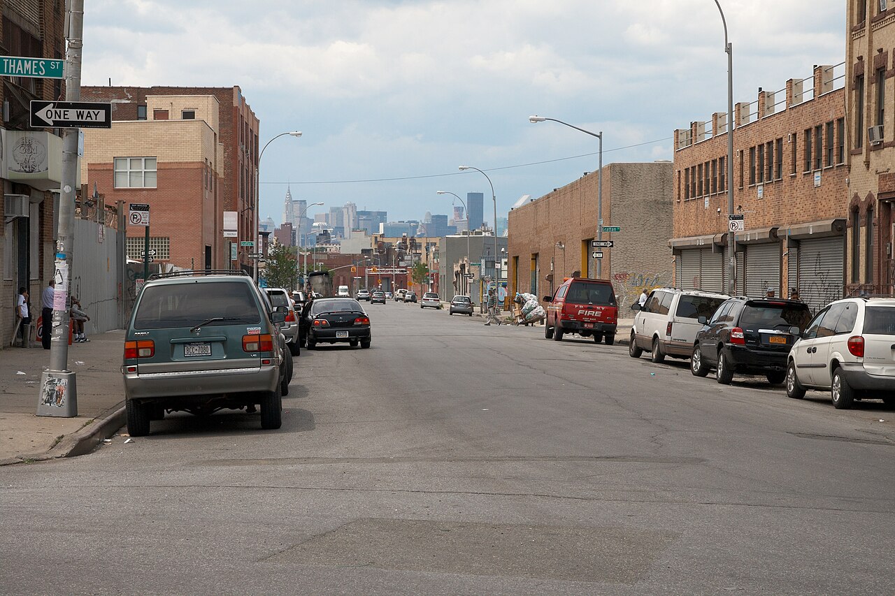 Knickerbocker Avenue at Thames Street, a wide low street of brick and cinder block buildings with roll down shutters, the Manhattan skyline closing the far end.