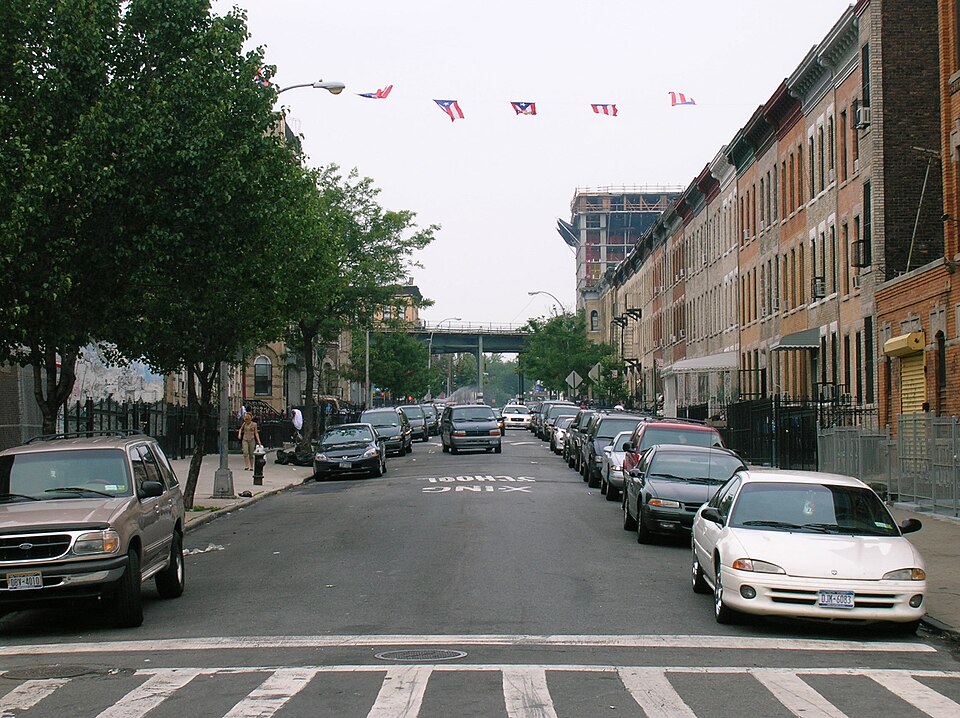 Knickerbocker Avenue looking down the block, brick rowhouses along the right, flags strung across the street and an elevated structure crossing in the distance.