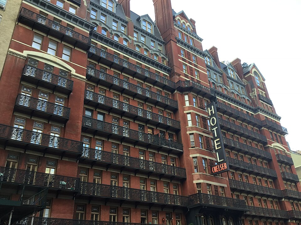 The Hotel Chelsea from West 23rd Street, red brick with iron balconies across every floor and the vertical hotel sign over the entrance.