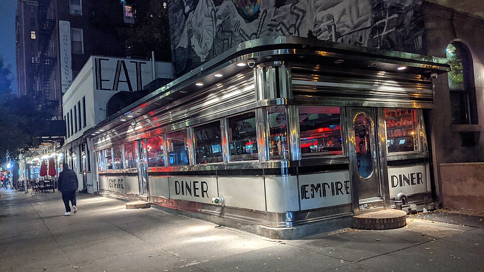 The Empire Diner at dusk, a stainless steel dining car on the corner with EAT painted in large letters on the wall behind it.