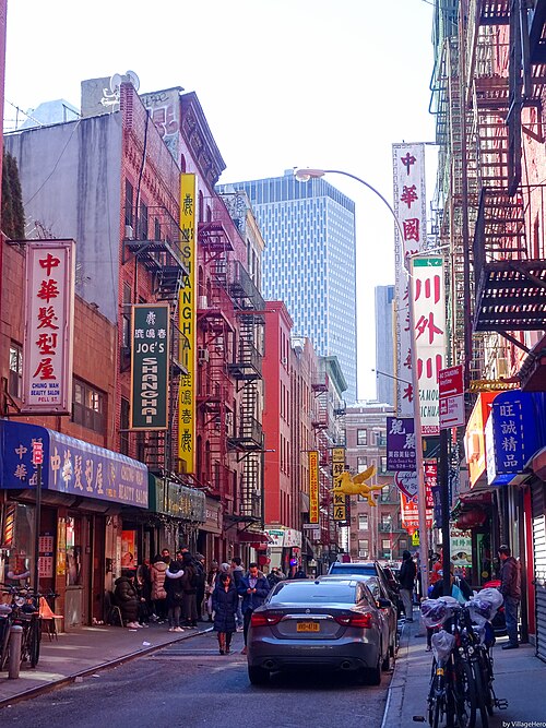 Pell Street looking east toward the Bowery, vertical signs and awnings down both sides, fire escapes above them and an office tower at the far end.