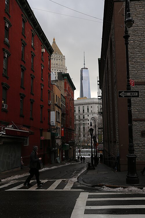 Mosco Street sloping west from Mott Street, red brick tenements on the left and the stone side wall of the Church of the Transfiguration on the right, with One World Trade Center and the gilded pyramid of the Foley Square courthouse beyond.