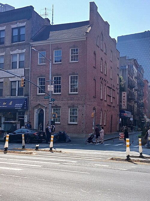 The Edward Mooney House at 18 Bowery on the corner of Pell Street, a three storey brick house with a pitched roof and dormer windows, seen across the Bowery on a sunny morning.