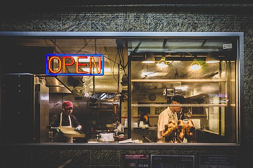 A restaurant kitchen seen through its window at night, roast ducks hanging on hooks under lamps, a neon open sign in the glass and two cooks working.