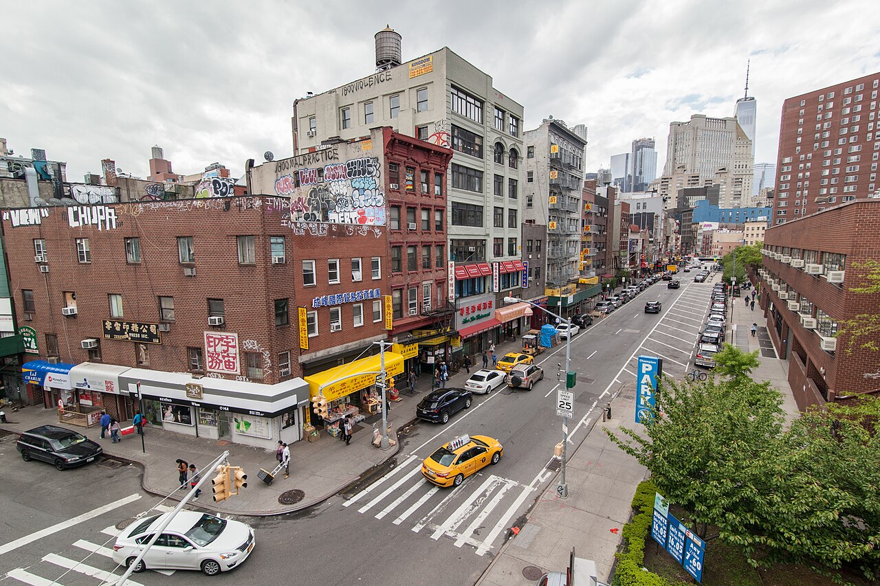 The corner of Division Street and Market Street from the walkway of the Manhattan Bridge, shopfronts with Chinese signs along the near side, a water tower on the roof above them and the Financial District towers in the distance.