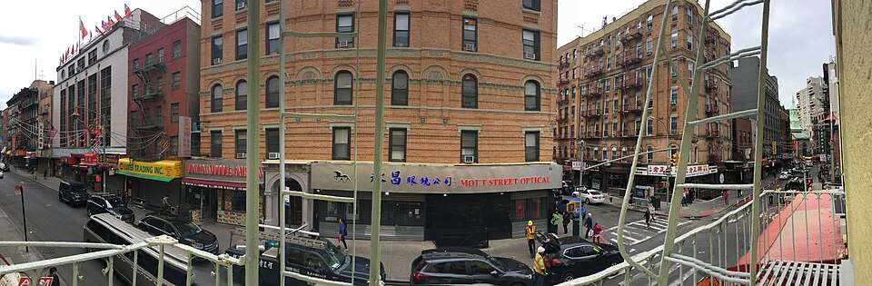 The corner of Mott Street and Bayard Street from a scaffold across the junction, a red brick tenement with an optician and a noodle shop under it, fire escapes above and shop signs down both streets.