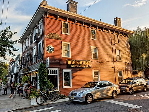 A three story clapboard corner building on the avenue in evening light, a restaurant at street level and people on a sidewalk bench.